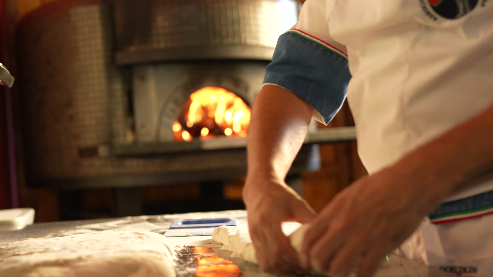 As mãos do pizzaiolo trabalhando a massa na bancada, com a boca do forno acesa atrás.