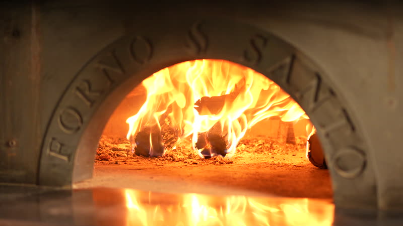 A boca do forno a lenha aceso, com as chamas altas e o reflexo do fogo no piso refratário.