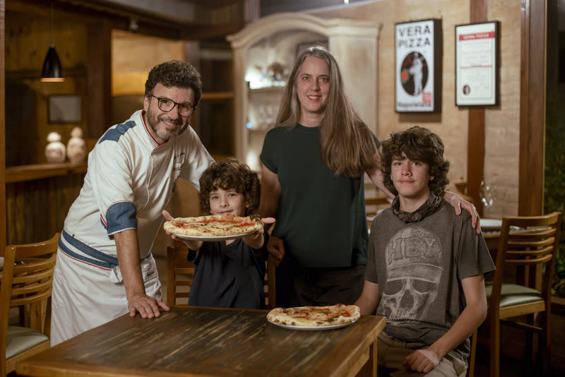 A família reunida no salão com uma pizza, sob as placas da Vera Pizza Napoletana.