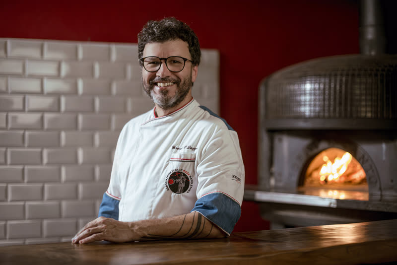 Gerardo Lazzari no balcão, com o forno a lenha aceso atrás dele.